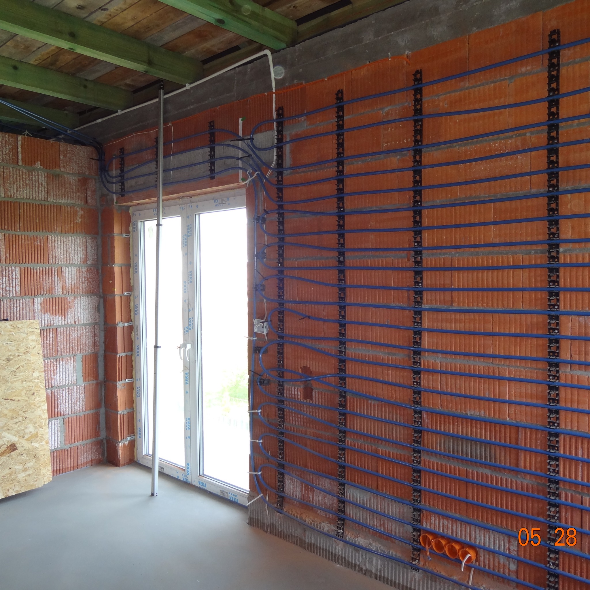 Instalacja ogrzewania ściennego z niebieskimi rurkami na ceglanej ścianie w surowym wnętrzu. Widoczne elementy montażowe i okno. Etap prac wykończeniowych.