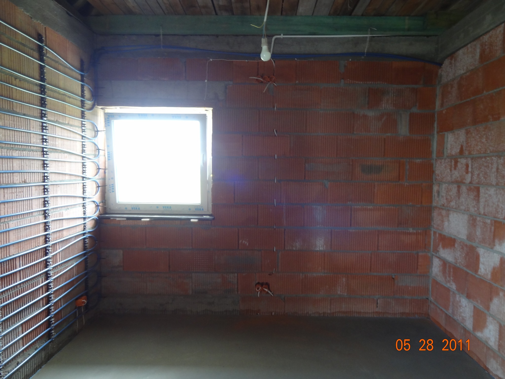 Instalacja ogrzewania podłogowego na ścianie z czerwonej cegły, widoczne okno i prowizoryczne oświetlenie. Etap prac przed wylaniem posadzki.