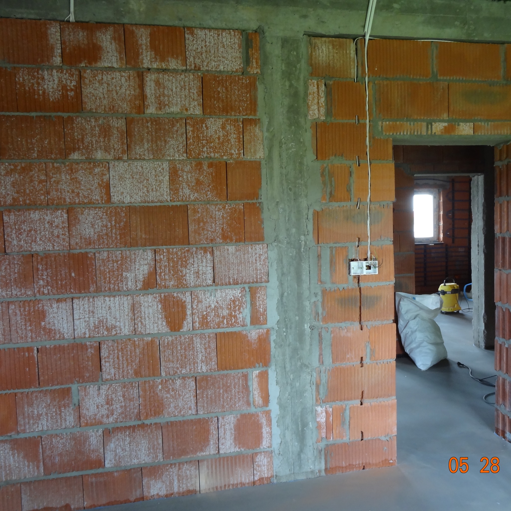 Wnętrze w trakcie budowy: ściany z czerwonej cegły, widoczne przewody elektryczne, gładka wylewka podłogowa, worek z materiałem i odkurzacz przemysłowy.