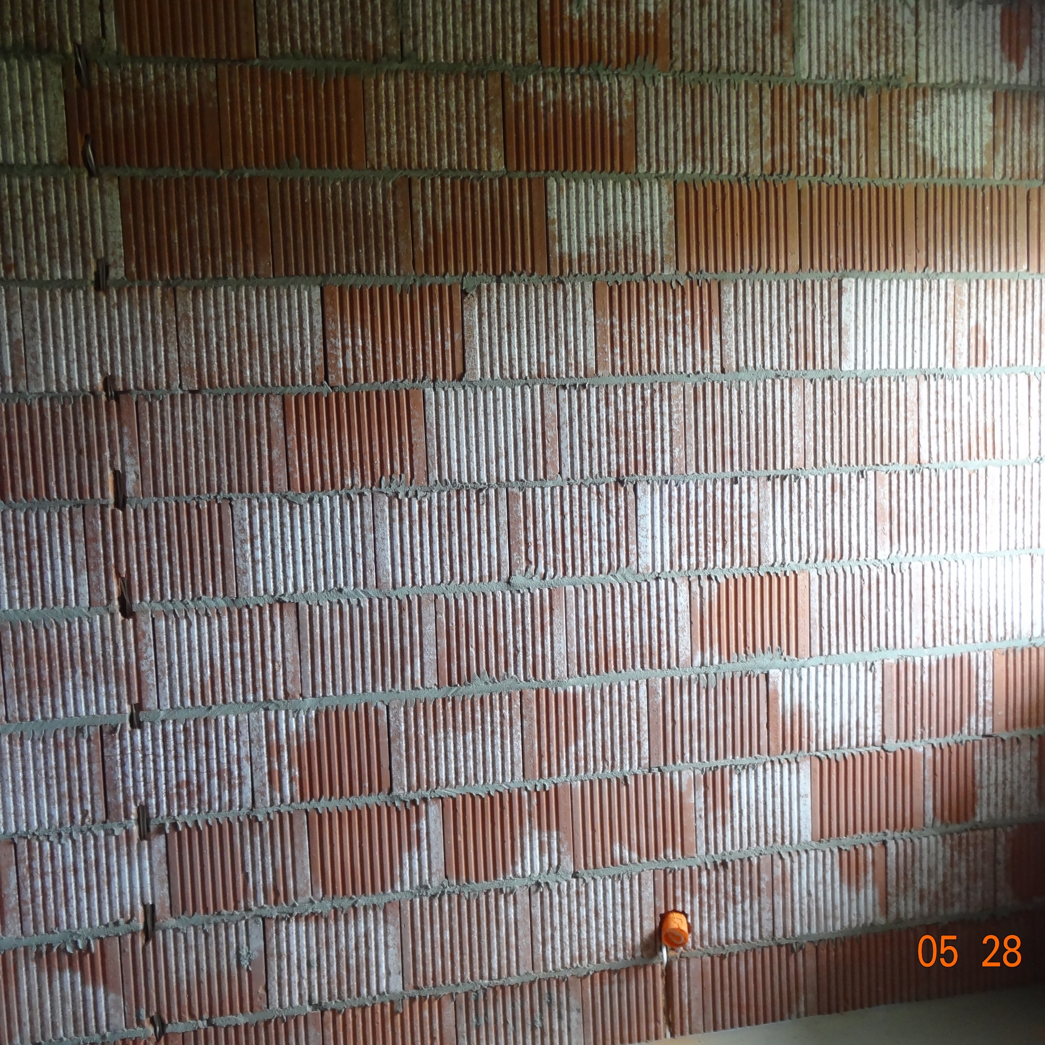 Ściana z czerwonej cegły dziurawki z widocznym otworem na puszkę elektryczną. Świeże spoiny między cegłami. Częściowo widoczne ślady zaprawy na powierzchni cegieł.