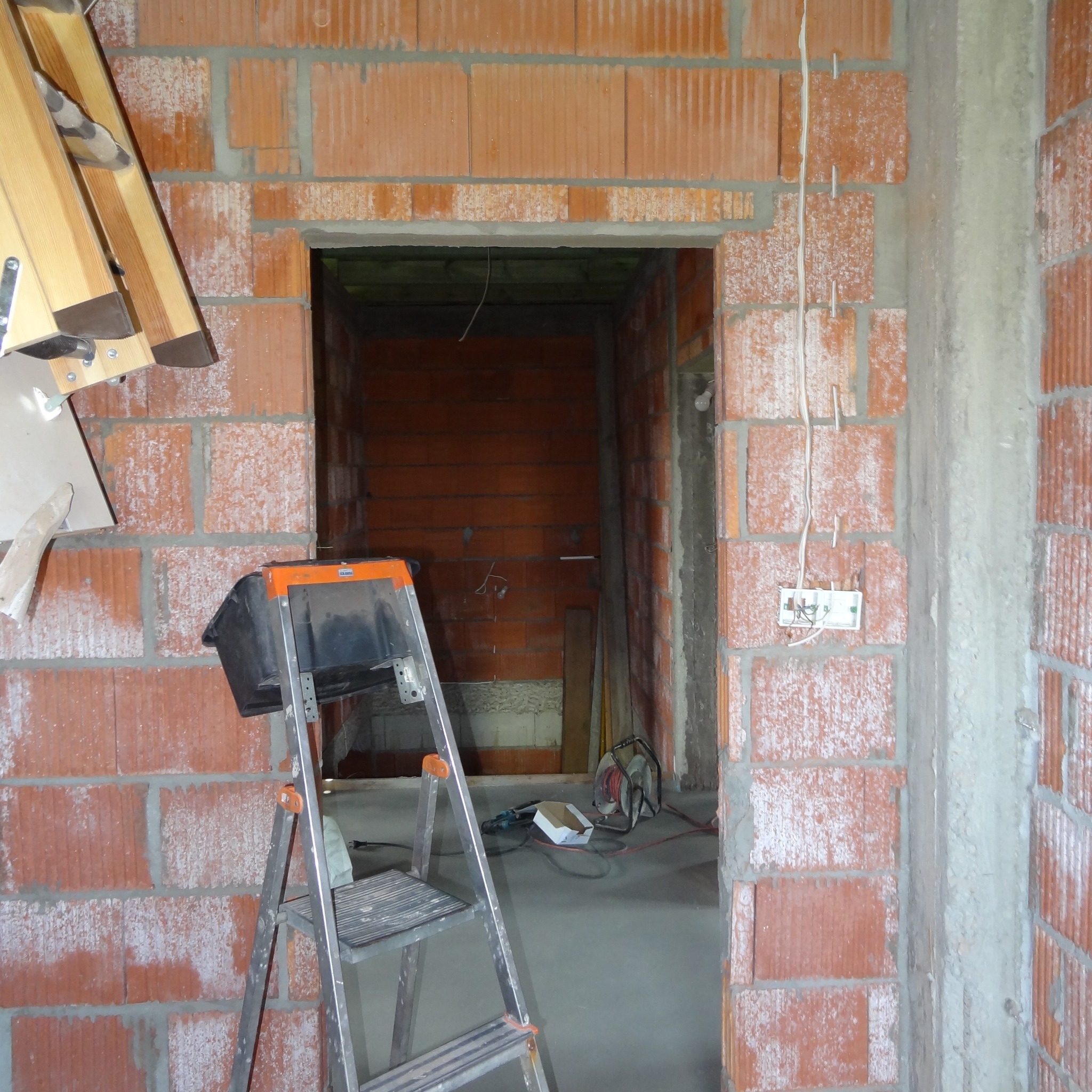 Wnętrze w trakcie prac murarskich z widoczną instalacją elektryczną; przewody wystają ze ściany. Na pierwszym planie drabina z kuwetą.