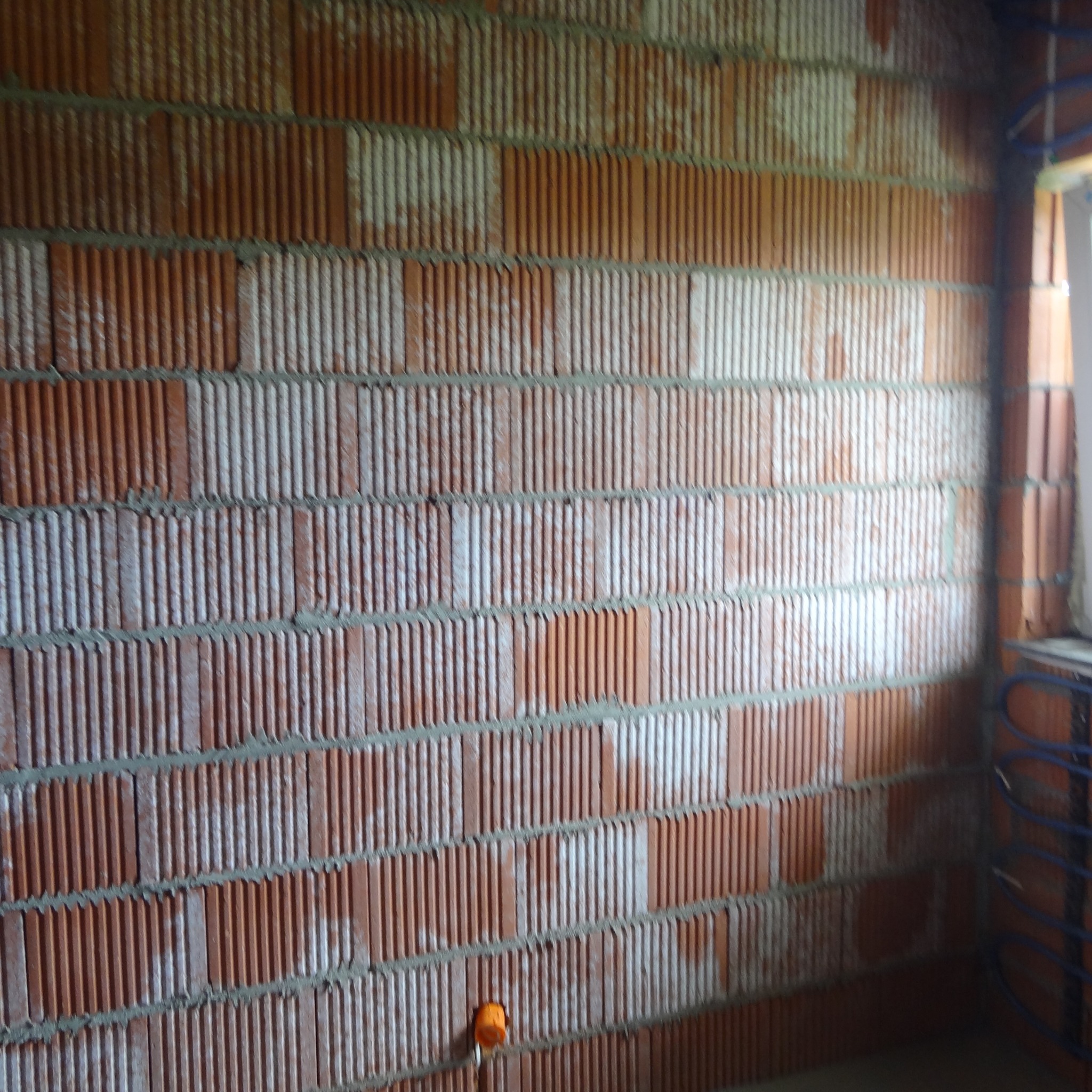 Ściana z czerwonej cegły dziurawki w trakcie budowy, widoczne ślady zaprawy. W rogu niebieskie rurki instalacji grzewczej i fragment okna. Pomarańczowa puszka elektryczna w ścianie.