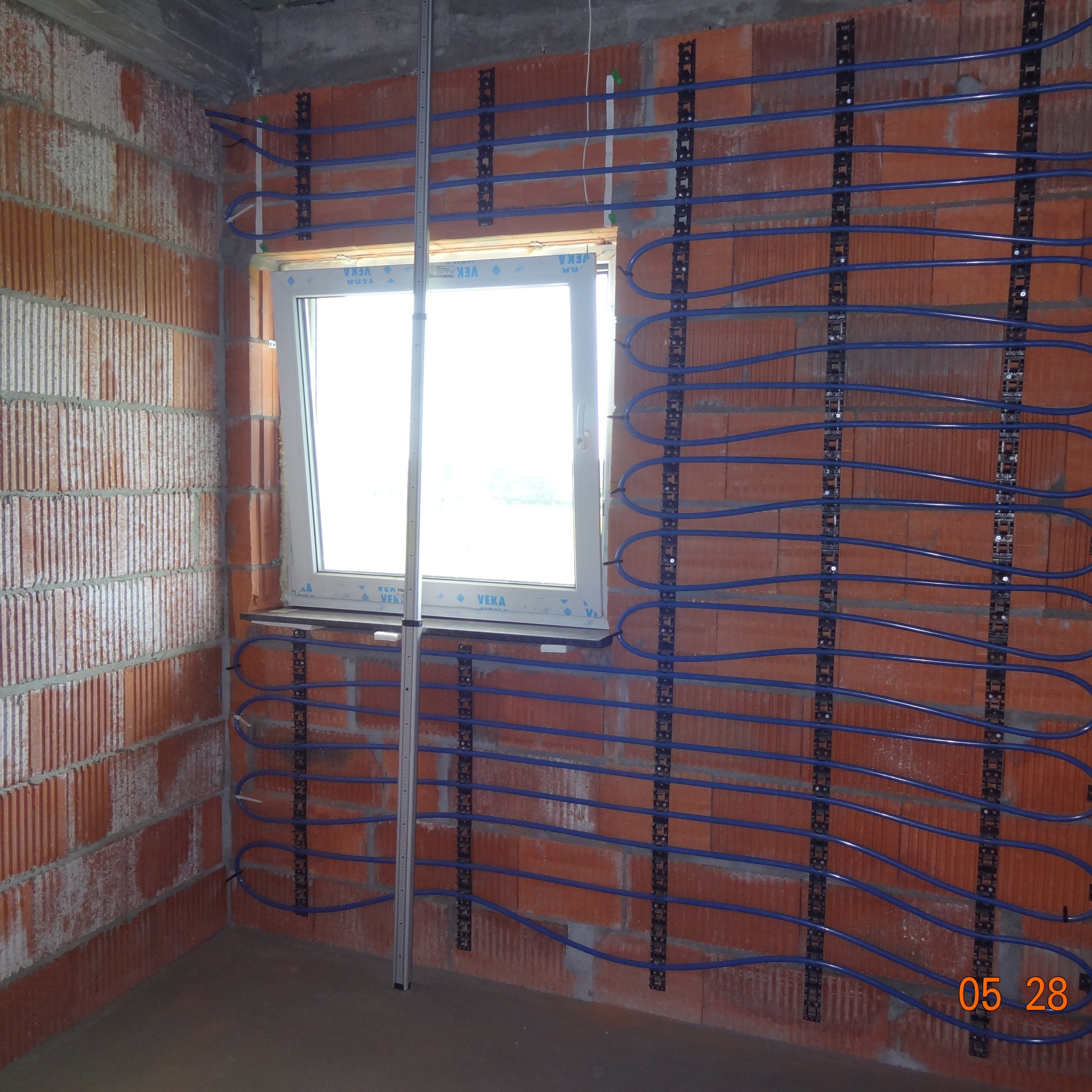 Ściana z cegły z zamontowanymi uchwytami i niebieskimi rurami, prawdopodobnie instalacja ogrzewania podłogowego. Widoczne okno i betonowa podłoga w surowym stanie.