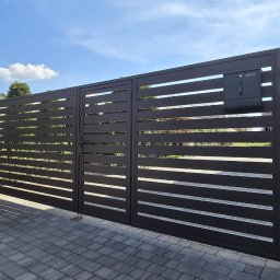 Bombowe Konstrukcje - Nowoczesna, pozioma brama metalowa w kolorze antracytowym z zamontowaną skrzynką na listy, na tle kostki brukowej i zieleni. Widok z zewnątrz posesji.