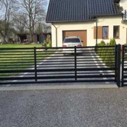 Nowoczesna brama wjazdowa z czarnych, poziomych profili, z automatyką i furtką. W tle dom z garażem i podjazdem z kostki. Widok z zewnątrz posesji, w tle zieleń.