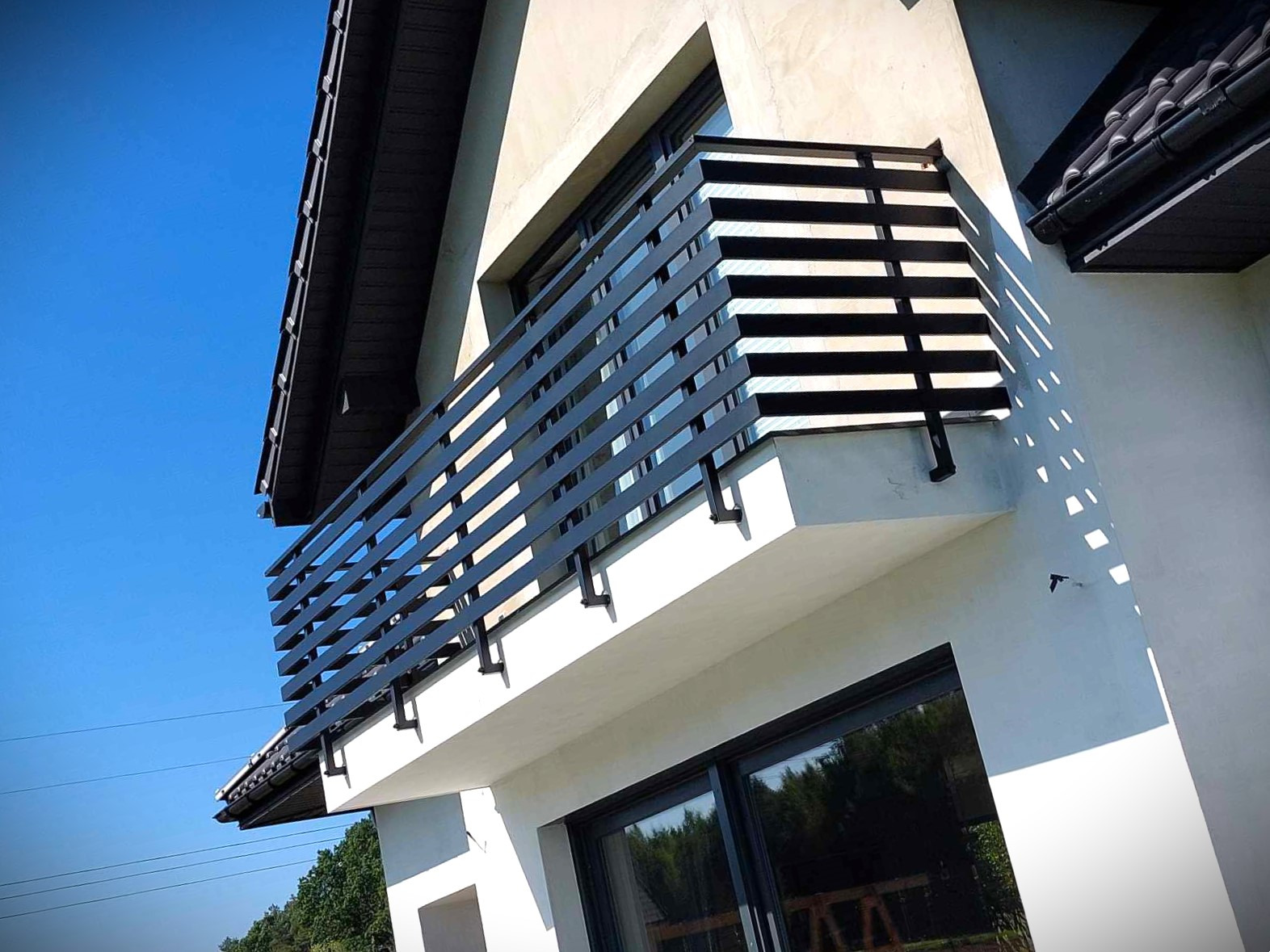 Nowoczesna, pozioma balustrada balkonowa z ciemnych, metalowych profili, zamontowana przy jasnym budynku. Ujęcie z dołu, widoczne niebo i okno.