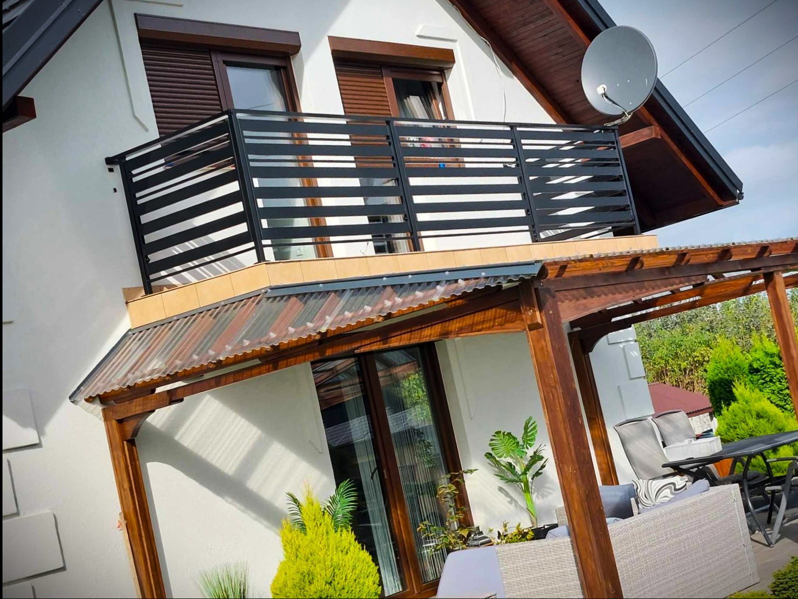 Nowoczesna, pozioma balustrada balkonowa w kolorze czarnym, z drewnianymi roletami w tle. Widoczna pergola z drewnianą konstrukcją i przezroczystym zadaszeniem.