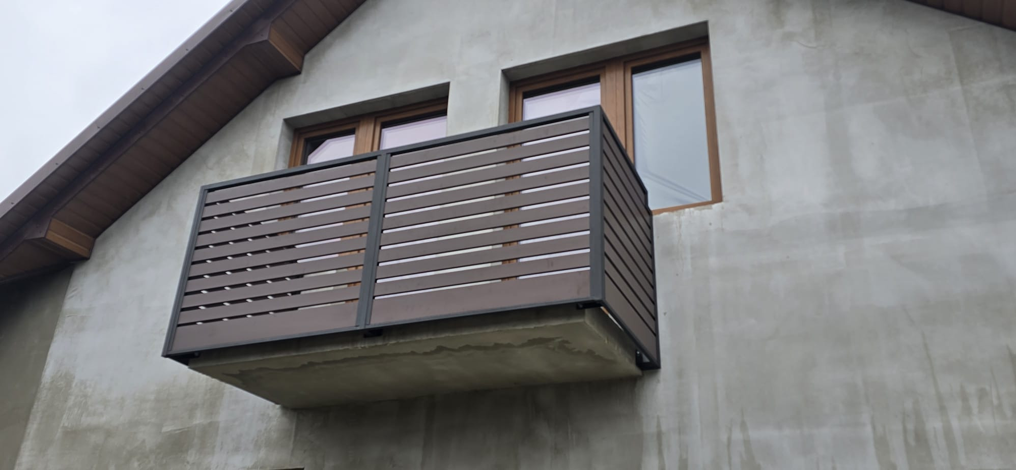 Balkon z poziomą, brązową balustradą z tworzywa sztucznego i ciemną ramą, zamontowany na szarym budynku. Widoczne okna z drewnianą ramą.