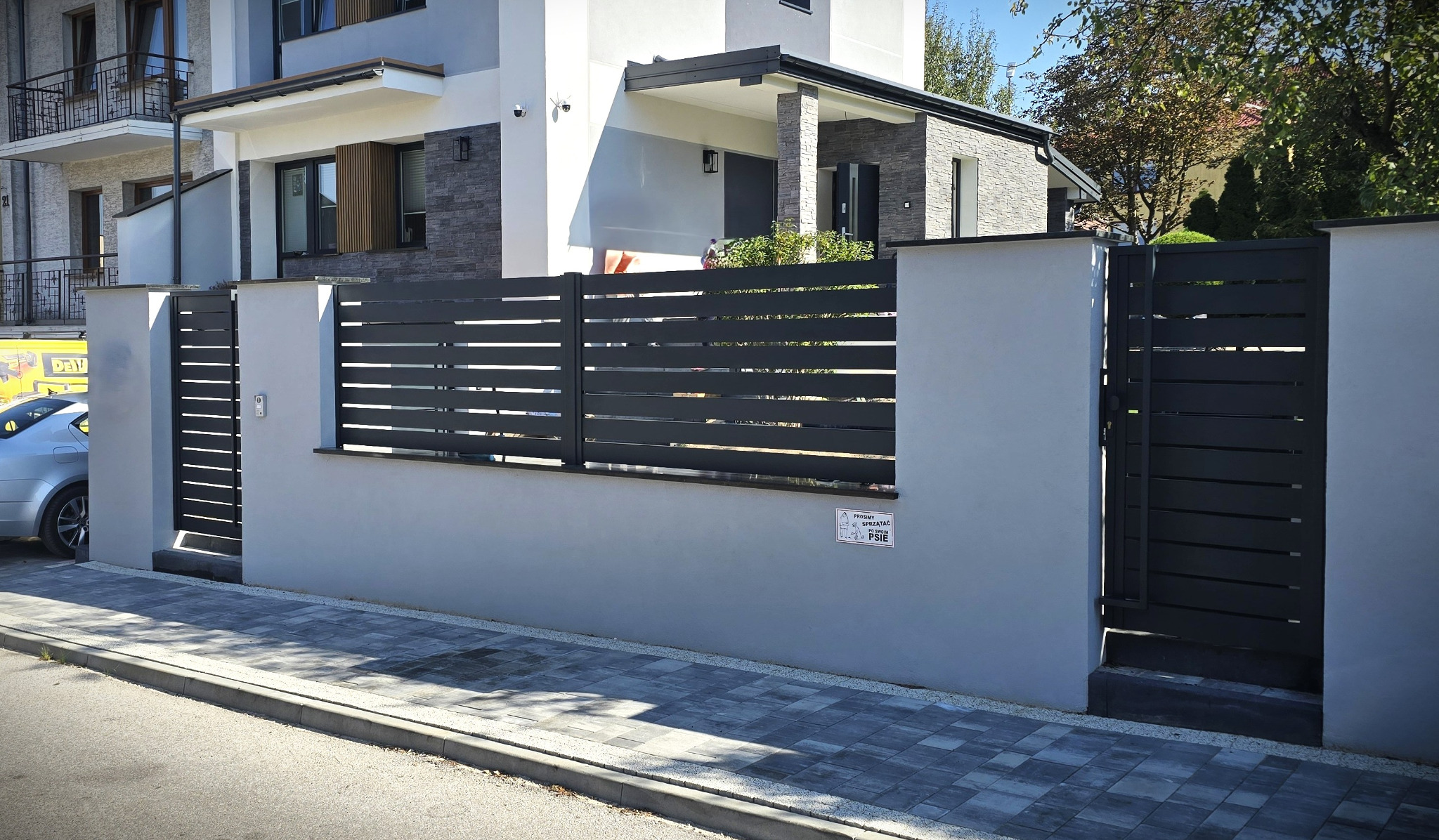 Nowoczesne ogrodzenie panelowe w kolorze antracytowym, z poziomymi przęsłami, wkomponowane w murki. Elegancki design pasujący do architektury domu.