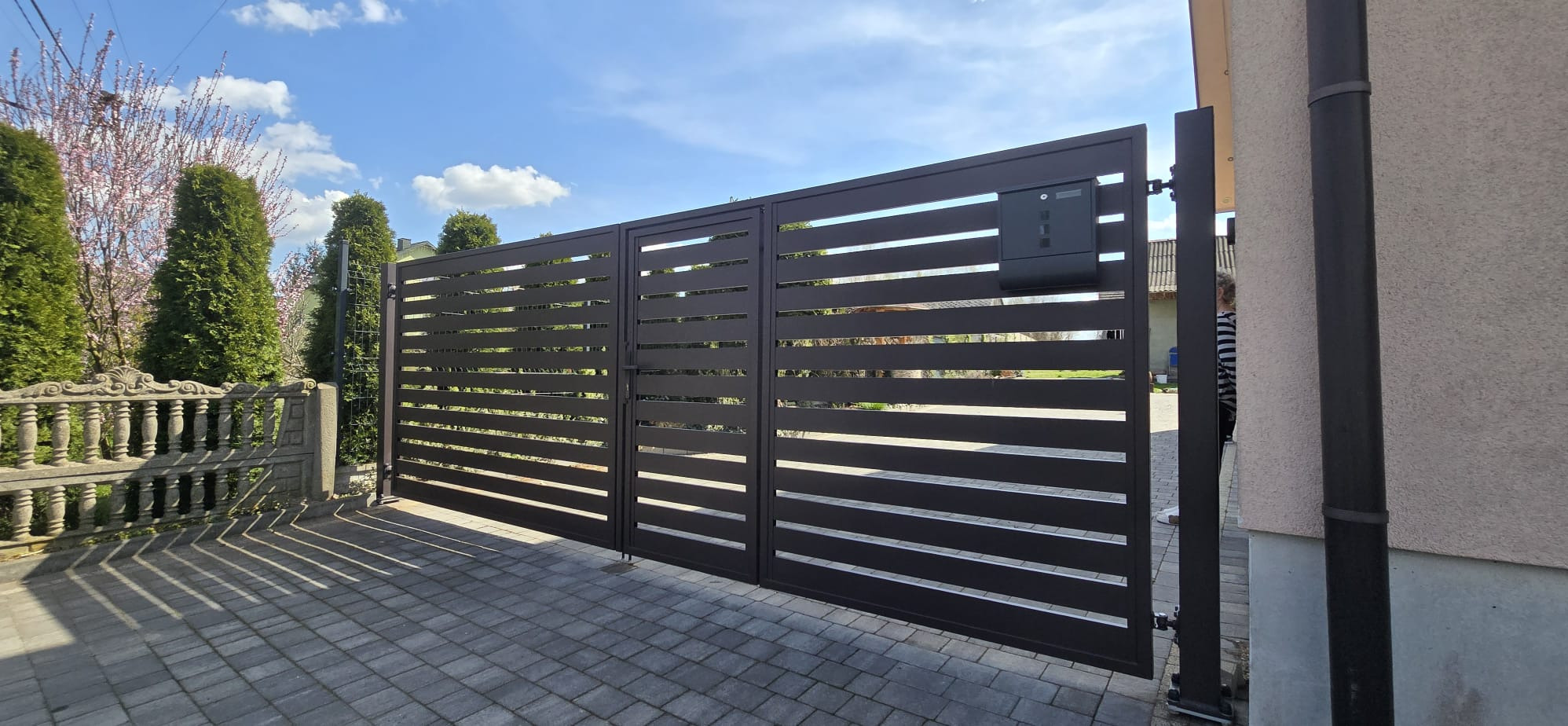 Nowoczesna, pozioma brama metalowa w kolorze antracytowym z zamontowaną skrzynką na listy, na tle kostki brukowej i zieleni. Widok z zewnątrz posesji.