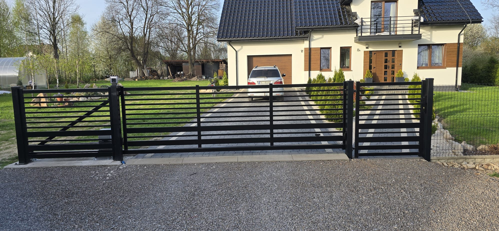 Nowoczesna brama wjazdowa z czarnych, poziomych profili, z automatyką i furtką. W tle dom z garażem i podjazdem z kostki. Widok z zewnątrz posesji, w tle zieleń.