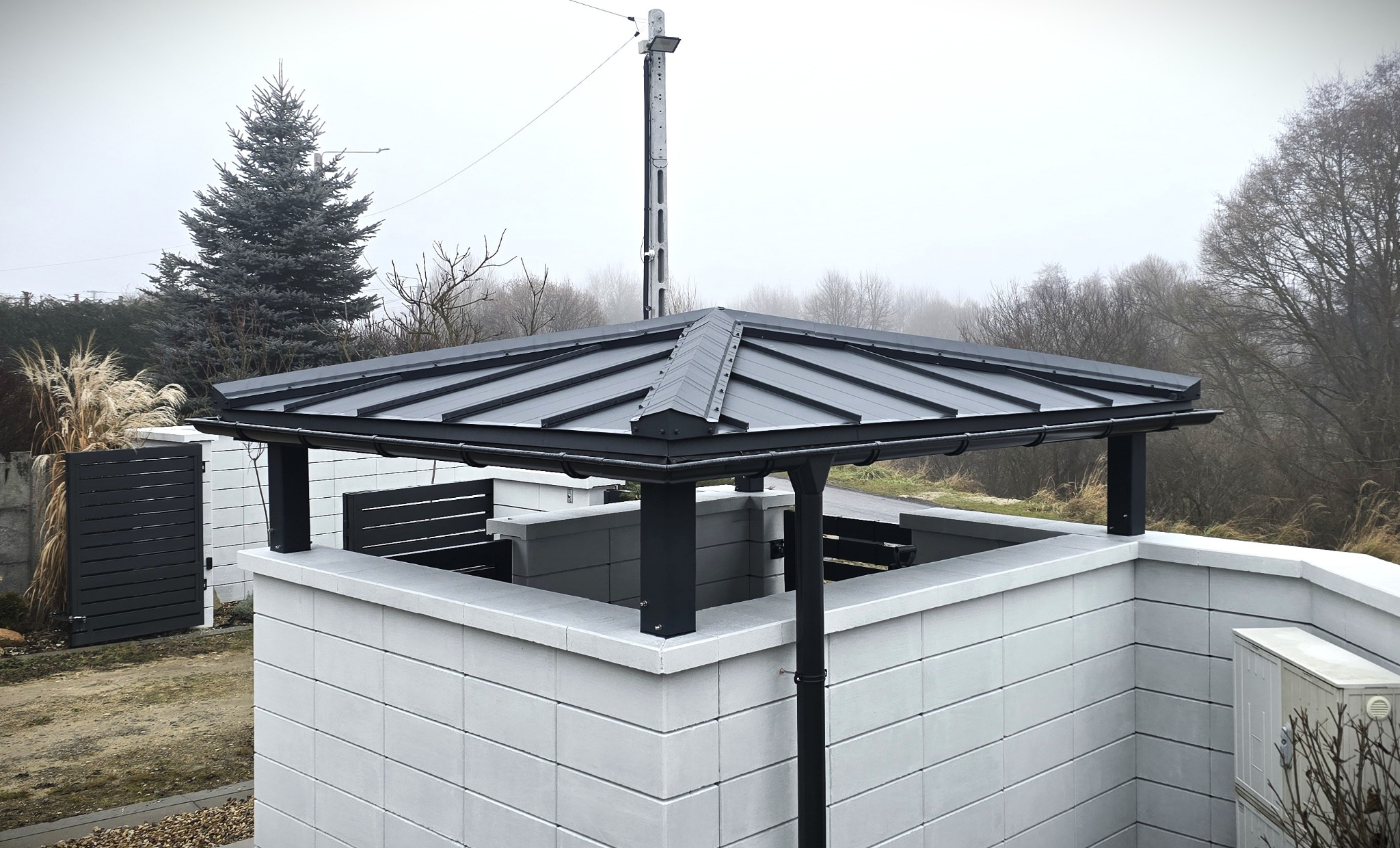 Nowoczesna pergola z szarej blachy trapezowej, wsparta na słupach i murze z betonowych bloczków. Minimalistyczny design, widok z góry, jesienna sceneria.