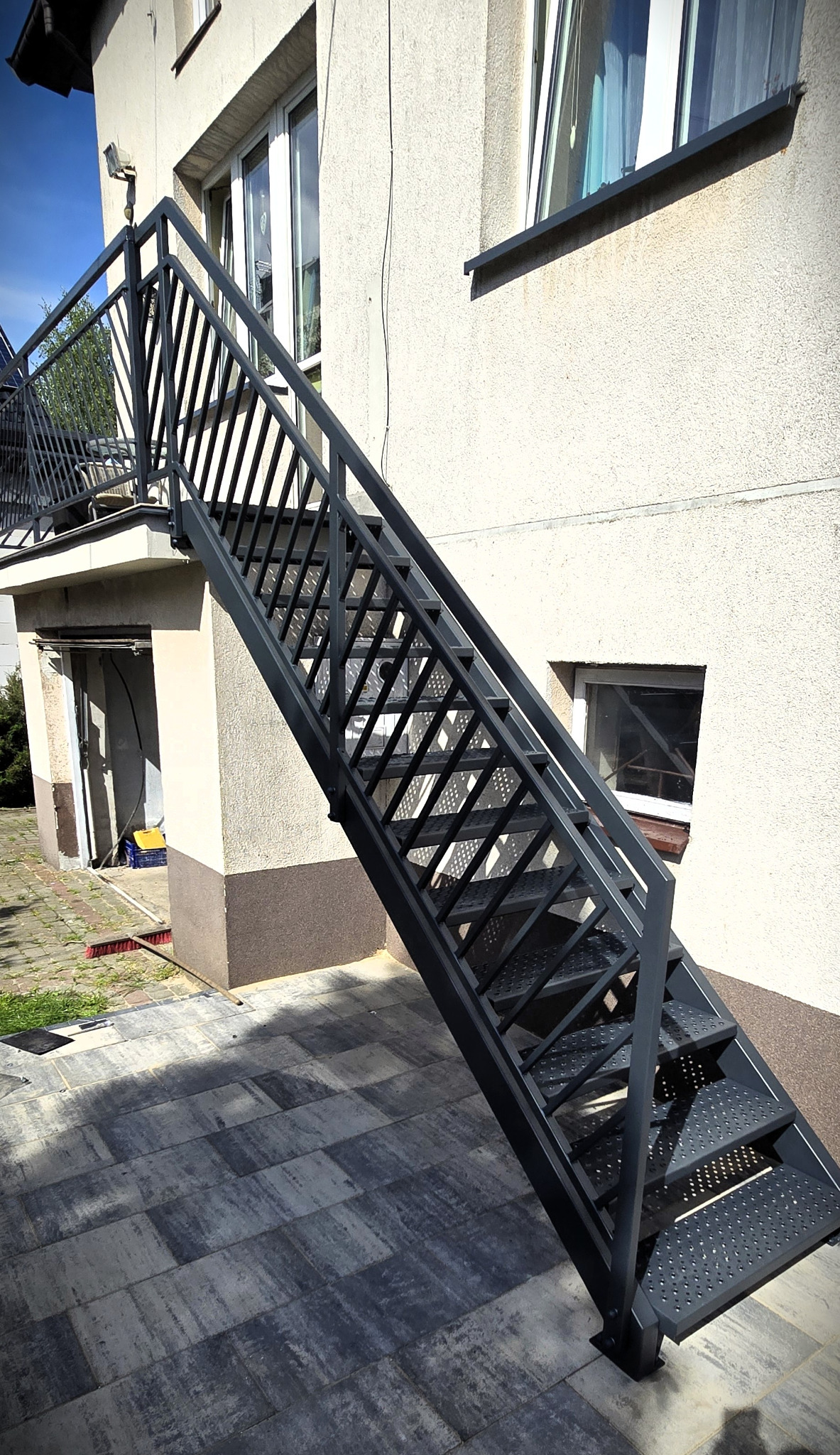 Zewnętrzne schody metalowe z ażurową balustradą, prowadzące na balkon. Szare schody na tle jasnej elewacji budynku i kostki brukowej.