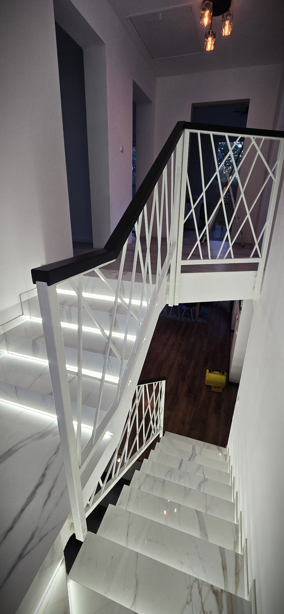 Nowoczesne schody z marmurowymi stopniami i białą balustradą z czarnym poręczą, oświetlone ledami, widziane z góry, z elementami wystroju wnętrza w tle.