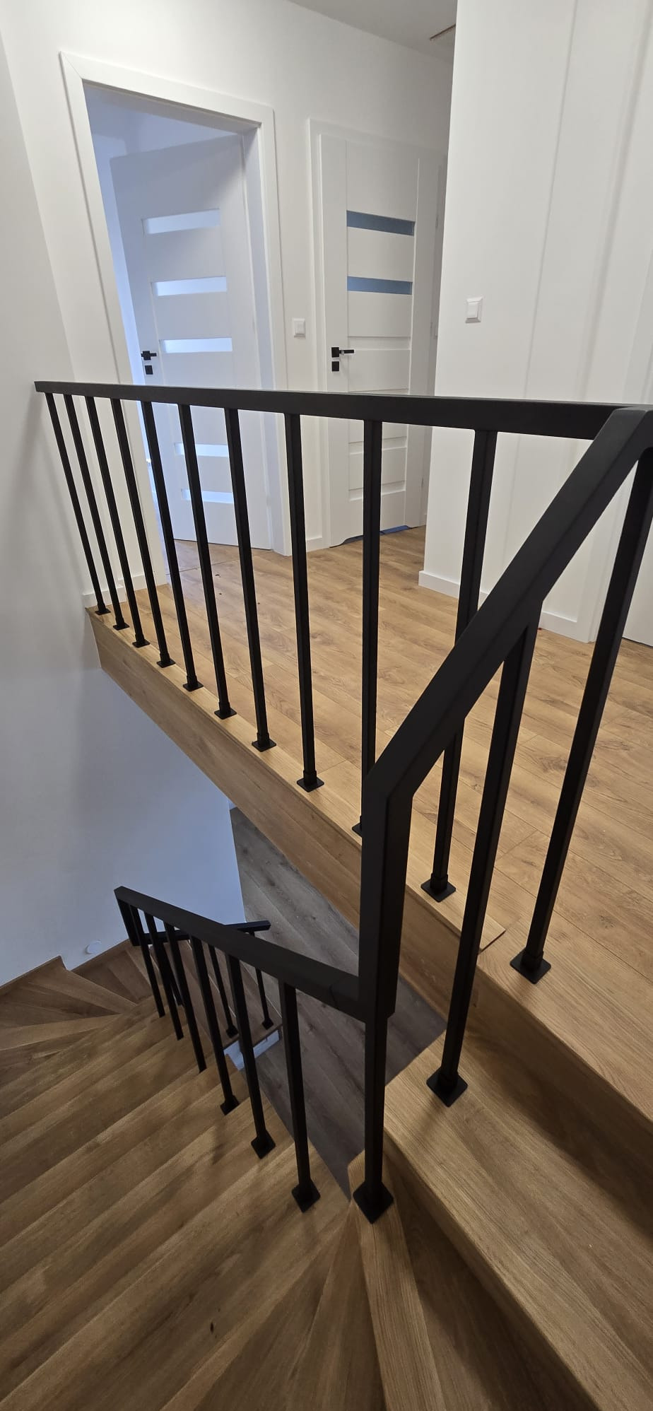 Nowoczesna balustrada metalowa w kolorze czarnym, z pionowymi słupkami i drewnianymi stopniami schodów. Widok z góry na jasne wnętrze z białymi drzwiami.