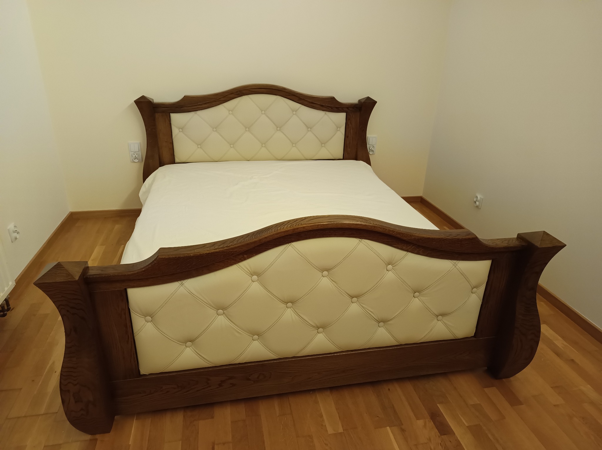 Drewniane łóżko z tapicerowanym, kremowym zagłówkiem w romby, widoczne w całości na tle parkietu i ściany w jasnym odcieniu.