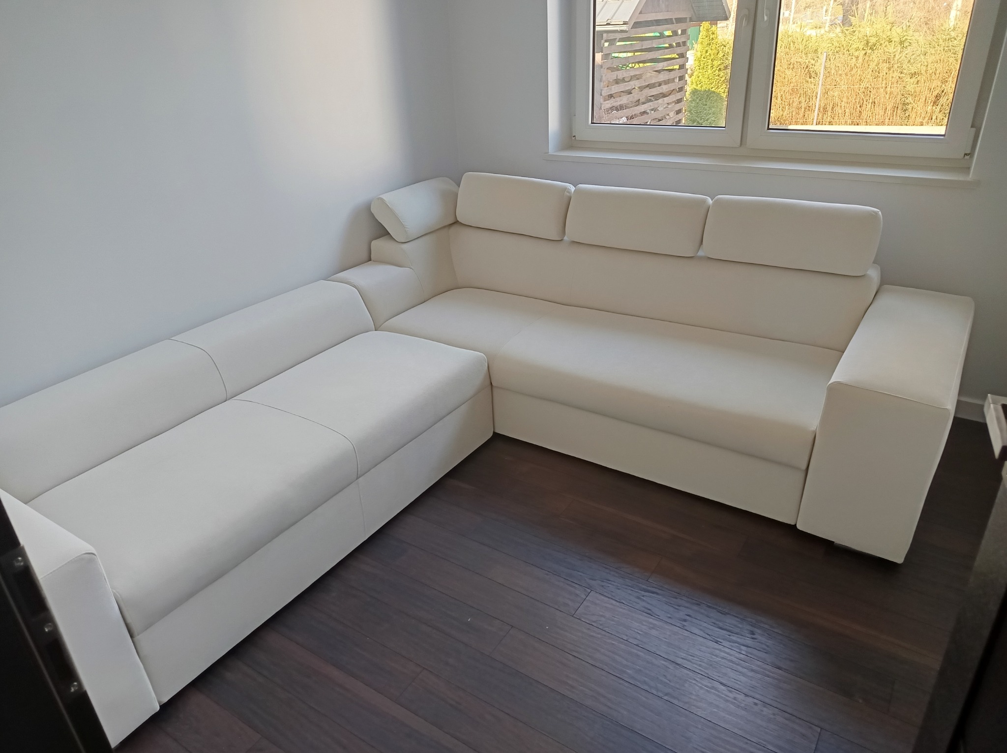 Biały, narożny zestaw wypoczynkowy z regulowanymi zagłówkami, ustawiony w rogu jasnego pokoju z drewnianą podłogą. Widok z góry.