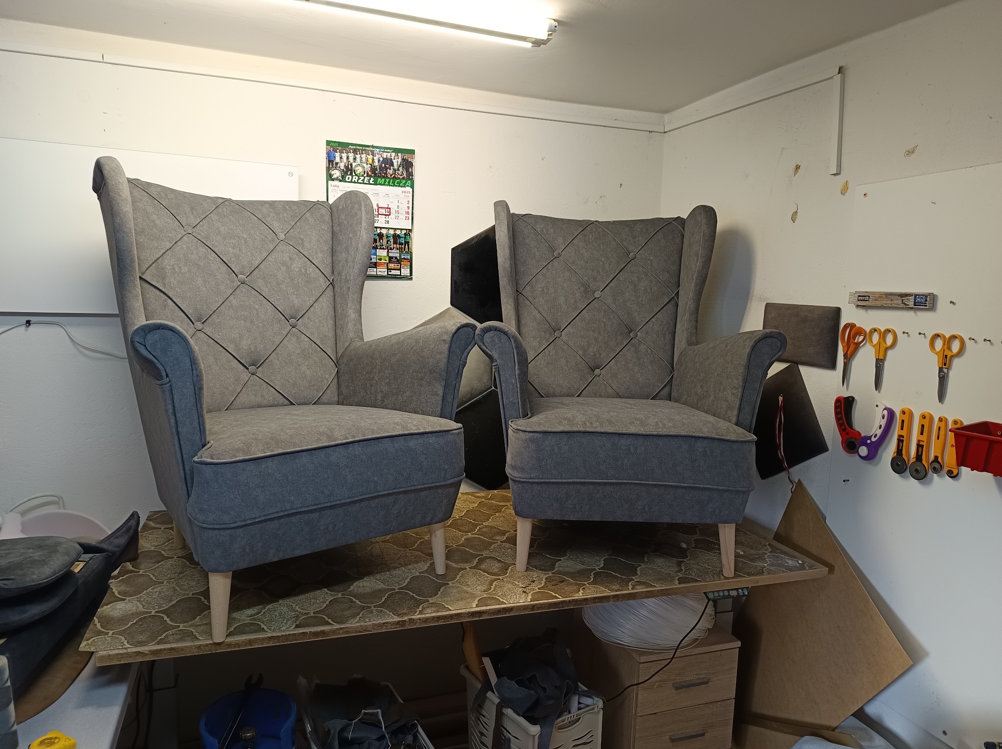 Dwa szare fotele typu uszak z pikowanym oparciem, ustawione na warsztatowym stole. W tle ściana z narzędziami tapicerskimi i kalendarzem.