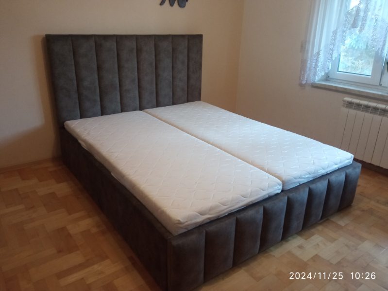 Tapicerowane łóżko z szarym wezgłowiem w pionowe pasy i dwoma białymi materacami, ustawione na drewnianej podłodze w pokoju z oknem.