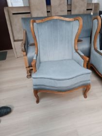 Błękitny fotel z drewnianą ramą na tle sofy, po renowacji tapicerskiej. Widoczne detale wykończenia i odnowiona tkanina. Jasna podłoga.