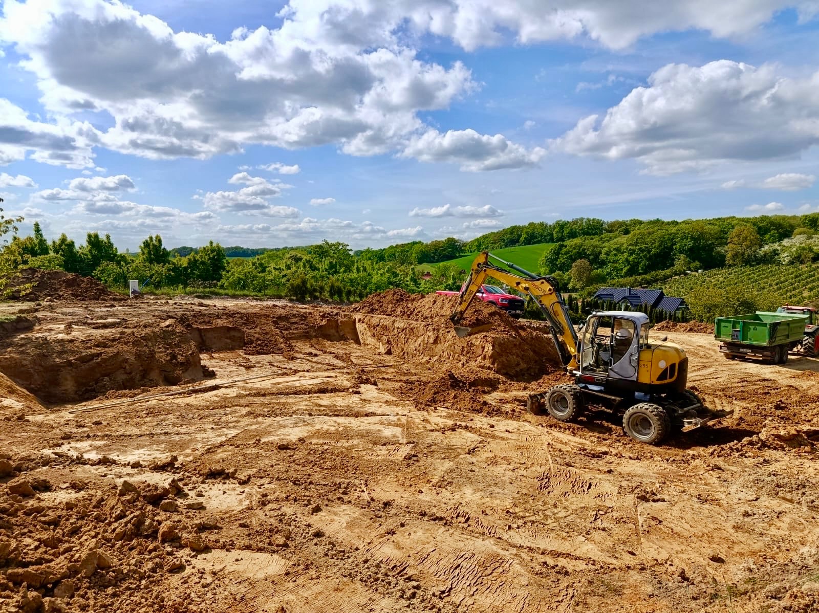 Żółta koparka na placu budowy, wykop pod fundamenty. W tle zielone drzewa i błękitne niebo z chmurami. Widoczny wywóz ziemi i przygotowanie terenu pod budowę.