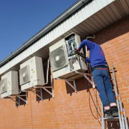 Serwisant na drabinie naprawia zewnętrzną jednostkę klimatyzacji Daikin, na ceglanej ścianie budynku. Widoczne trzy klimatyzatory, instalacja elektryczna i narzędzia.