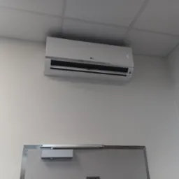 Biała klimatyzacja LG, zamontowana na ścianie nad drzwiami w pomieszczeniu z kasetonowym sufitem. Widoczny wyświetlacz temperatury i logo producenta.