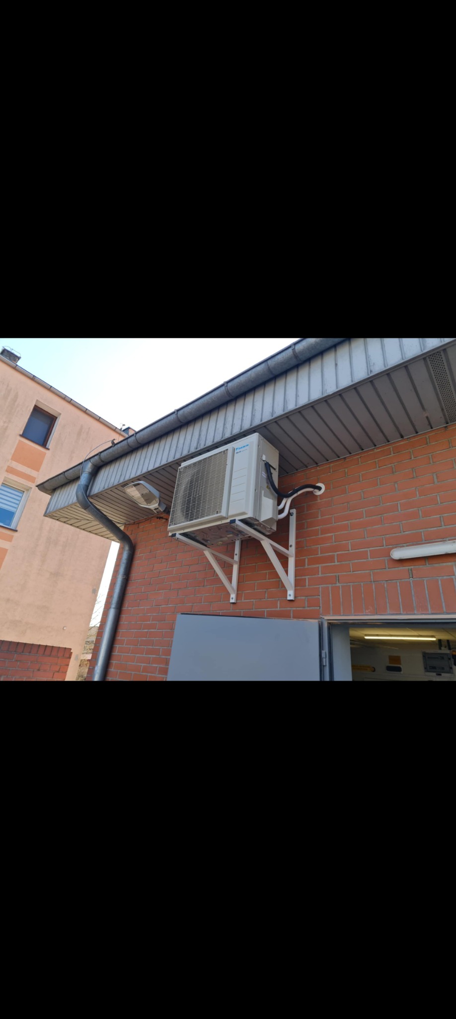 Zamontowany klimatyzator Daikin na ceglanej ścianie budynku. Widoczne białe wsporniki i rury. Częściowo widoczny budynek sąsiedni i fragment dachu.
