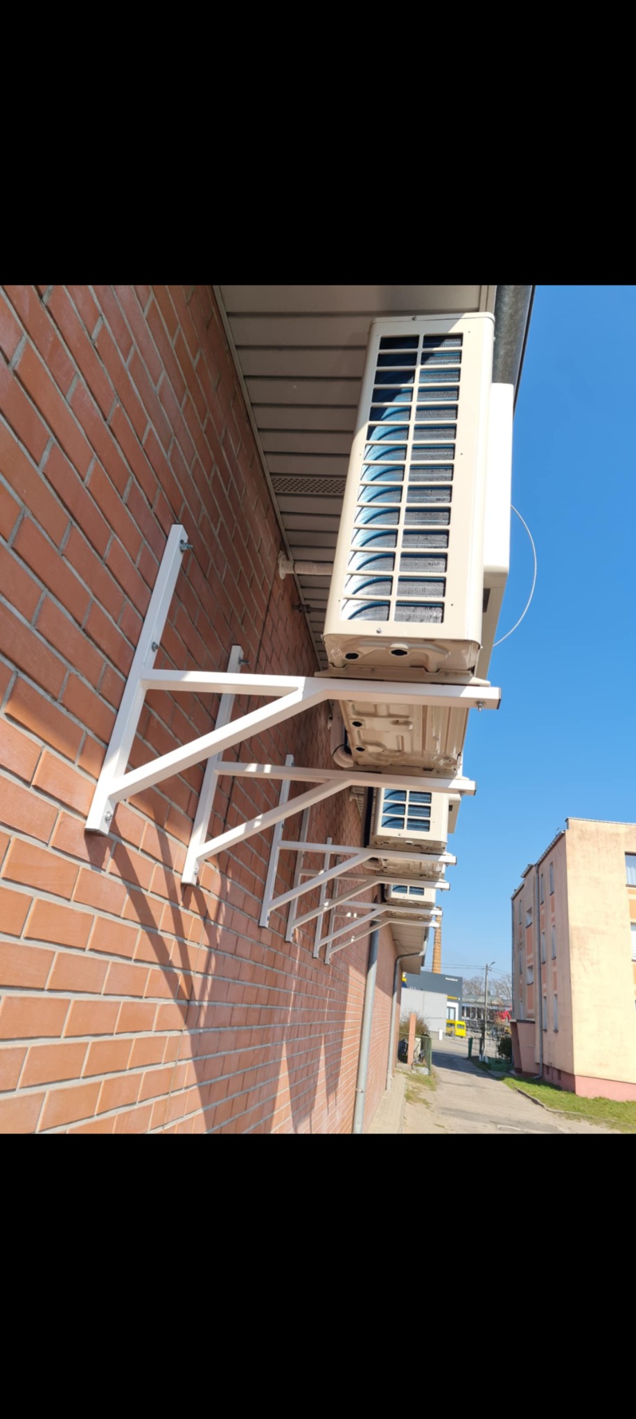 Zewnętrzne jednostki klimatyzacji zamontowane na ścianie z cegły, wsparte na białych, metalowych wspornikach. Widok z boku, w tle budynek i błękitne niebo.