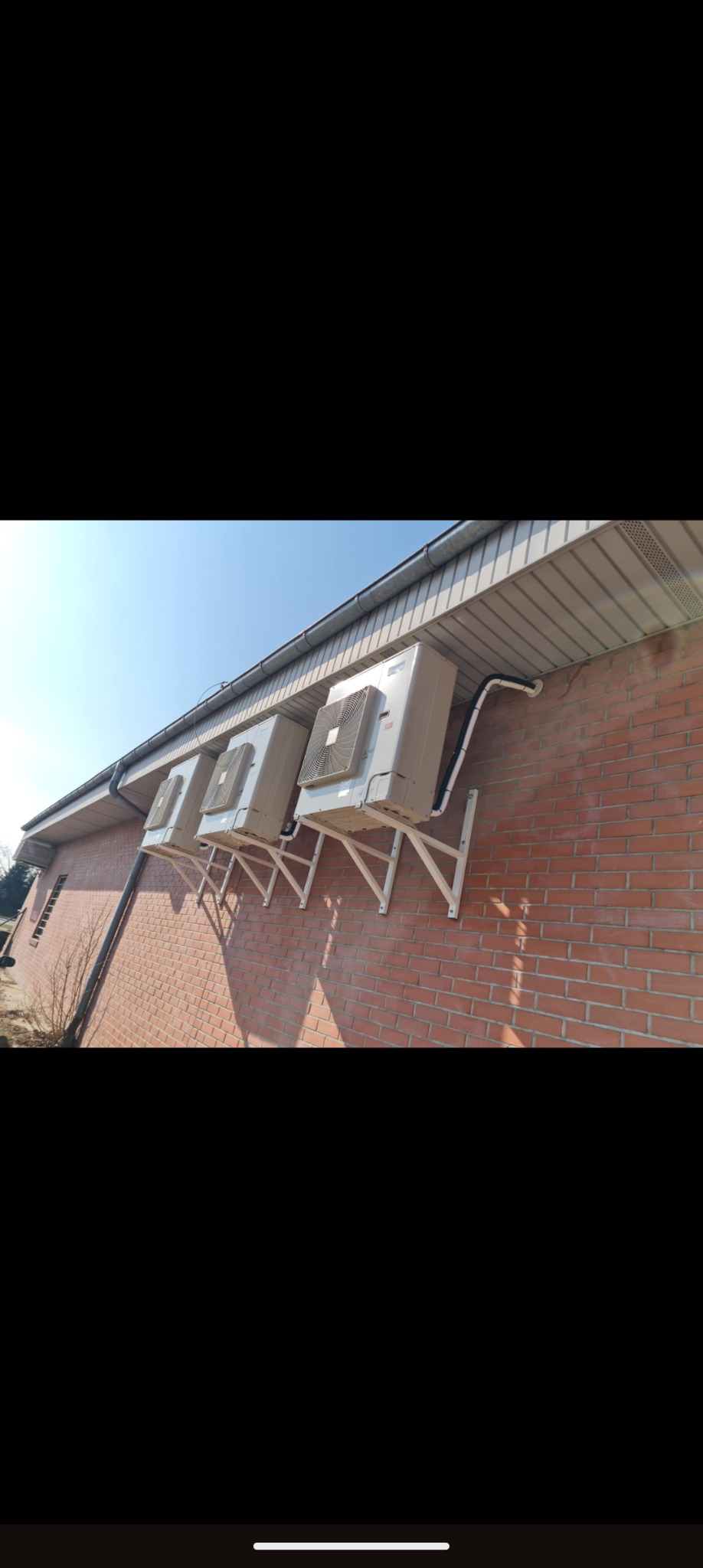 Trzy klimatyzatory zamontowane na ceglanej ścianie budynku, wsparte na białych, metalowych wspornikach. Widoczne rury odprowadzające. Słoneczny dzień.