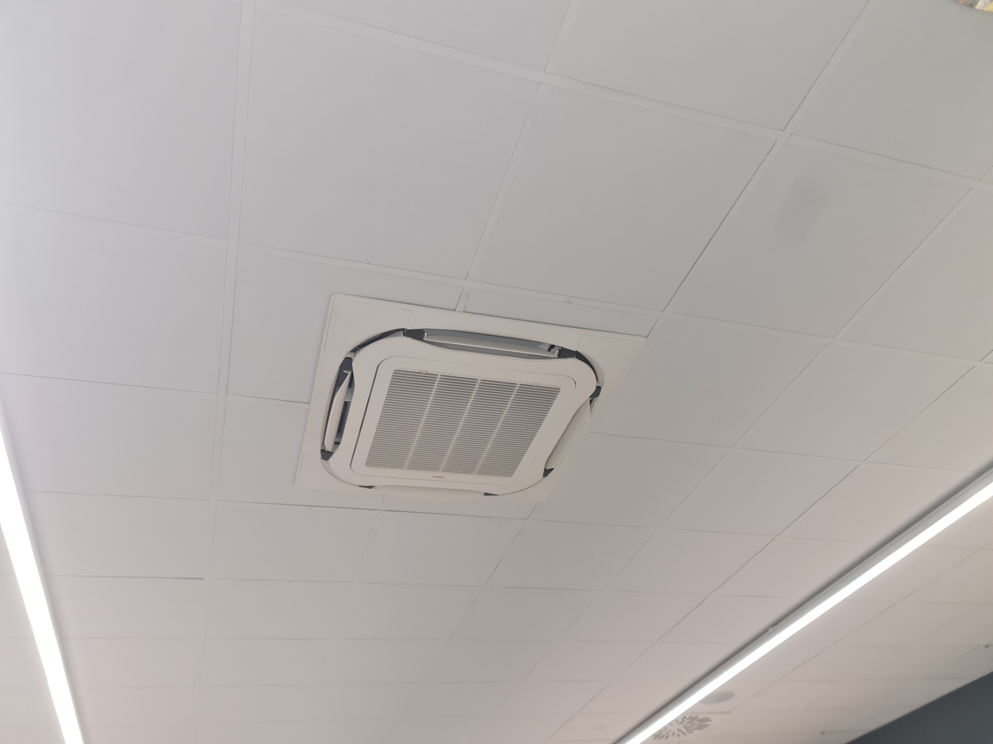 Kasetonowa klimatyzacja sufitowa wkomponowana w biały sufit podwieszany, widoczna z dołu pod kątem. Obok lampa liniowa LED.