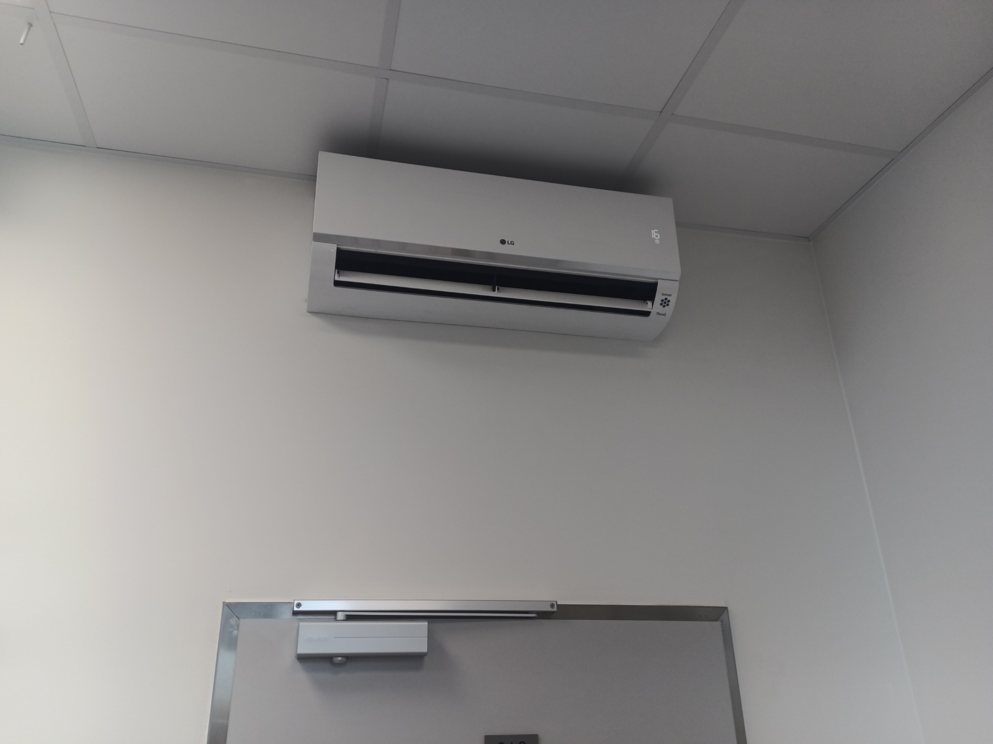 Biała klimatyzacja LG, zamontowana na ścianie nad drzwiami w pomieszczeniu z kasetonowym sufitem. Widoczny wyświetlacz temperatury i logo producenta.