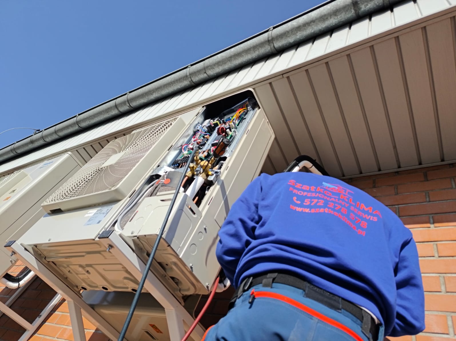 Serwisant w niebieskiej bluzie naprawia zewnętrzną jednostkę klimatyzacji na wsporniku, widoczne przewody i instalacja, ceglana ściana budynku w tle.