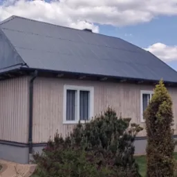 Dom z nowym, szarym dachem z blachy falistej w Myślenicach. Jasna elewacja drewniana, białe okna. W tle błękitne niebo z obłokami i sąsiednie budynki.