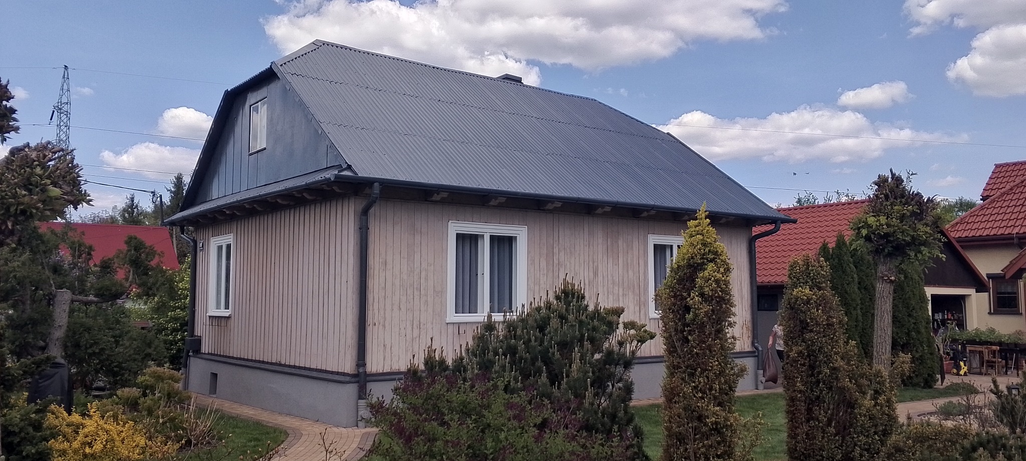 Dom z nowym, szarym dachem z blachy falistej w Myślenicach. Jasna elewacja drewniana, białe okna. W tle błękitne niebo z obłokami i sąsiednie budynki.