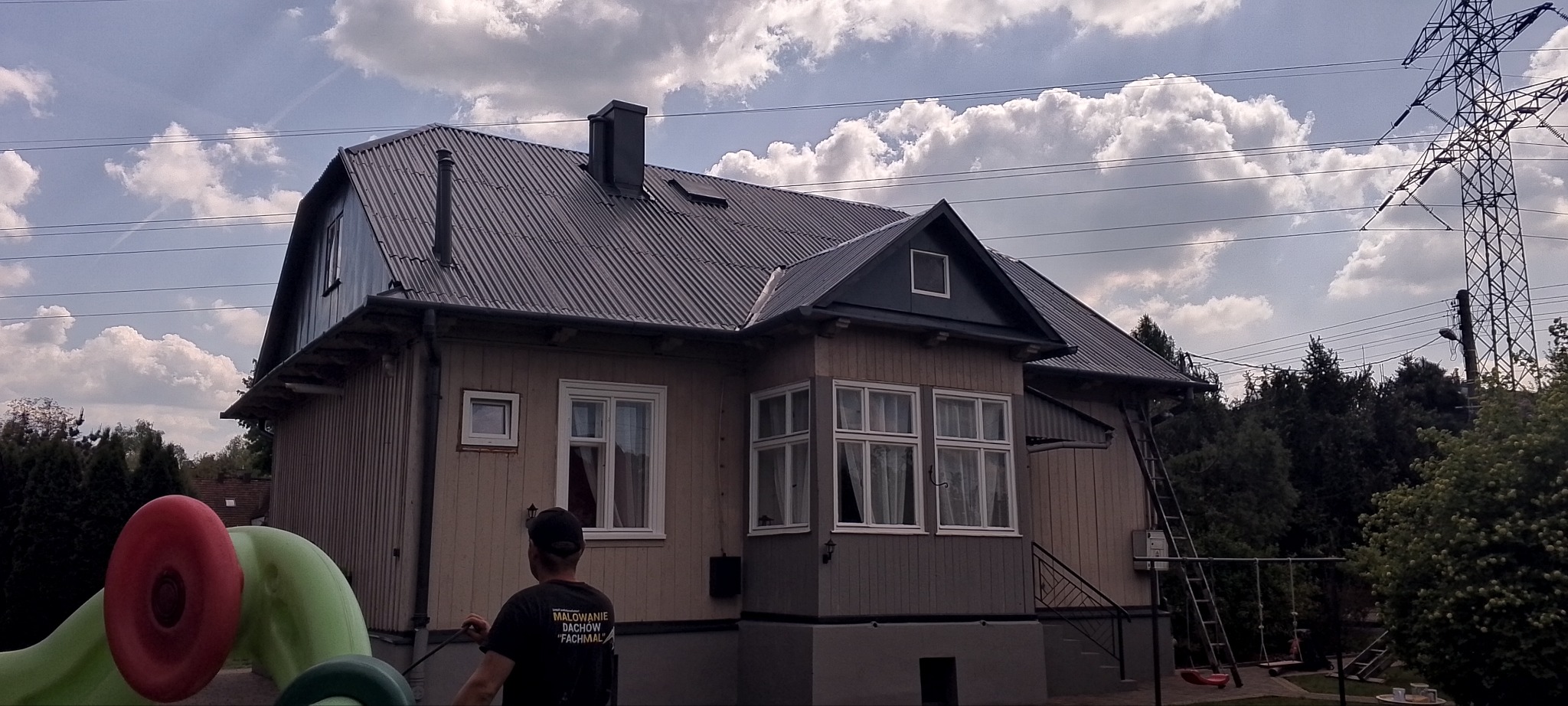 Dom z pomalowanym dachem i elewacją w odcieniach szarości i beżu, drabina oparta o ścianę, widoczna osoba w koszulce z napisem o malowaniu dachów.