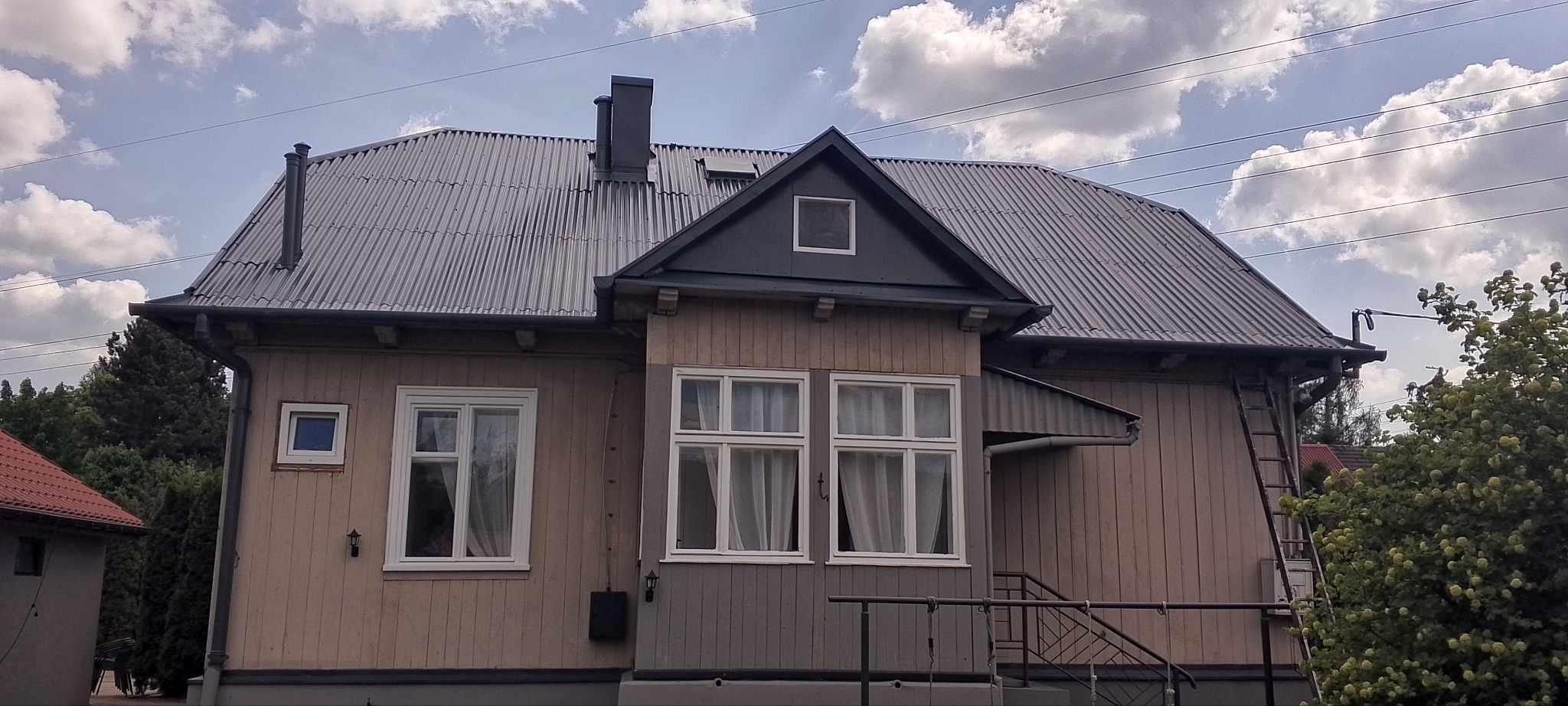 Odnowiony dom z blachy trapezowej w szarym kolorze. Widoczne okna z białymi ramami, drabina oparta o rynnę, błękitne niebo z chmurami w tle.