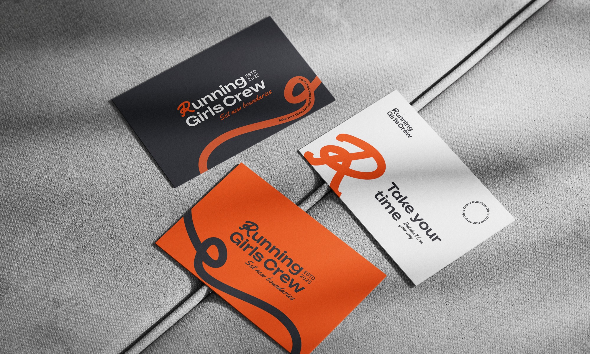 Trzy wizytówki Running Girls Crew w kolorach pomarańczowym, czarnym i białym, leżące na szarej, teksturowanej powierzchni. Minimalistyczny design z logo i hasłem.
