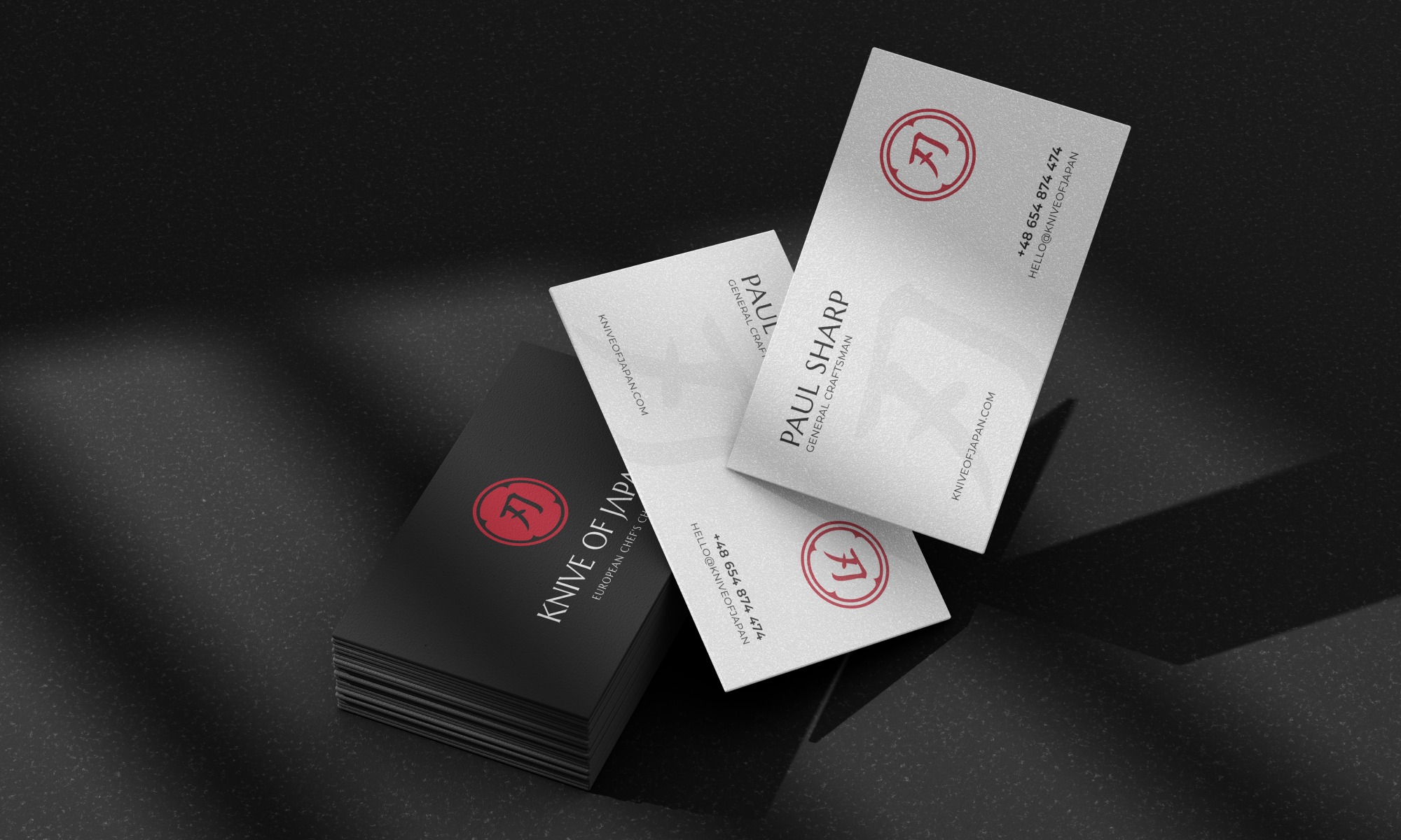 Stos czarnych wizytówek 'Knife of Japan' i dwie białe wizytówki 'Paul Sharp' na ciemnym tle. Minimalistyczny design, logo z symbolem japońskiego ostrza. Eleganckie i profesjonalne.