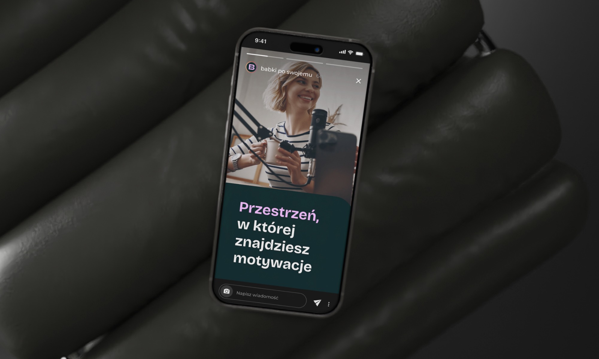 Ekran smartfona w dłoni, prezentujący relację z wizerunkiem kobiety przed mikrofonem i tekstem 'Przestrzeń, w której znajdziesz motywacje'.