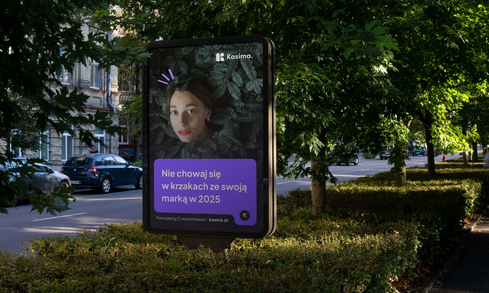 Billboard z wizerunkiem kobiety na tle roślin i tekstem 'Nie chowaj się w krzakach ze swoją marką w 2025'. Reklama marki Kasima na tle zieleni miejskiej.