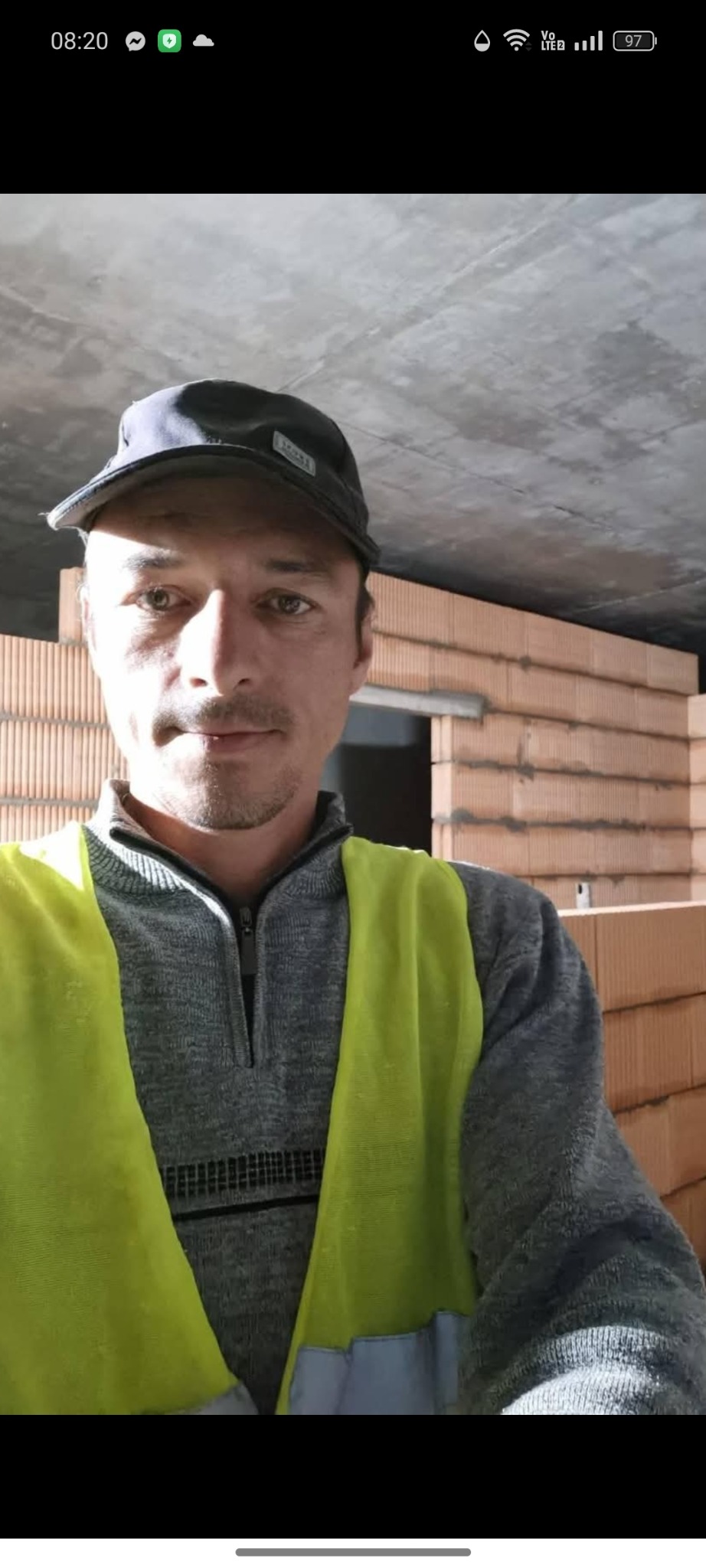 Mężczyzna w kamizelce odblaskowej i czapce na tle surowych ścian z czerwonej cegły w trakcie budowy domu. Widoczny betonowy strop.