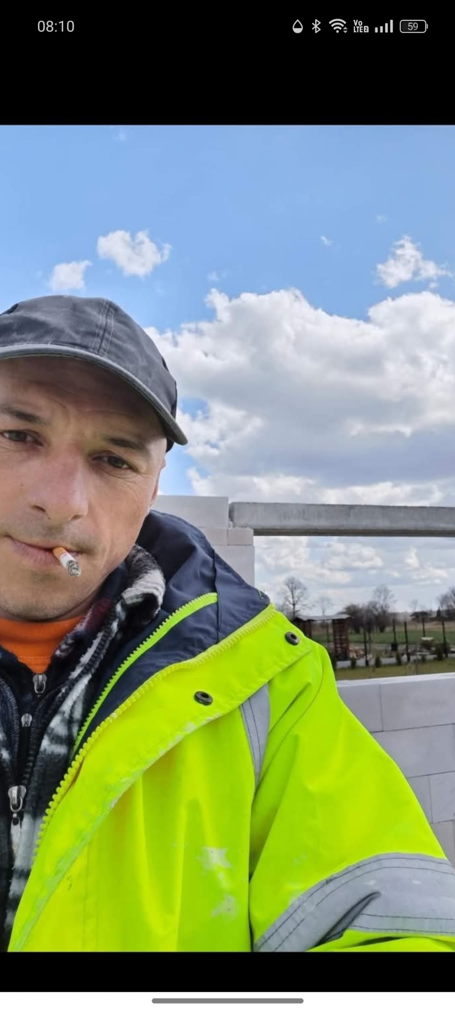 Mężczyzna w odblaskowej kurtce i czapce z papierosem na tle budowy z białych bloczków i błękitnego nieba.