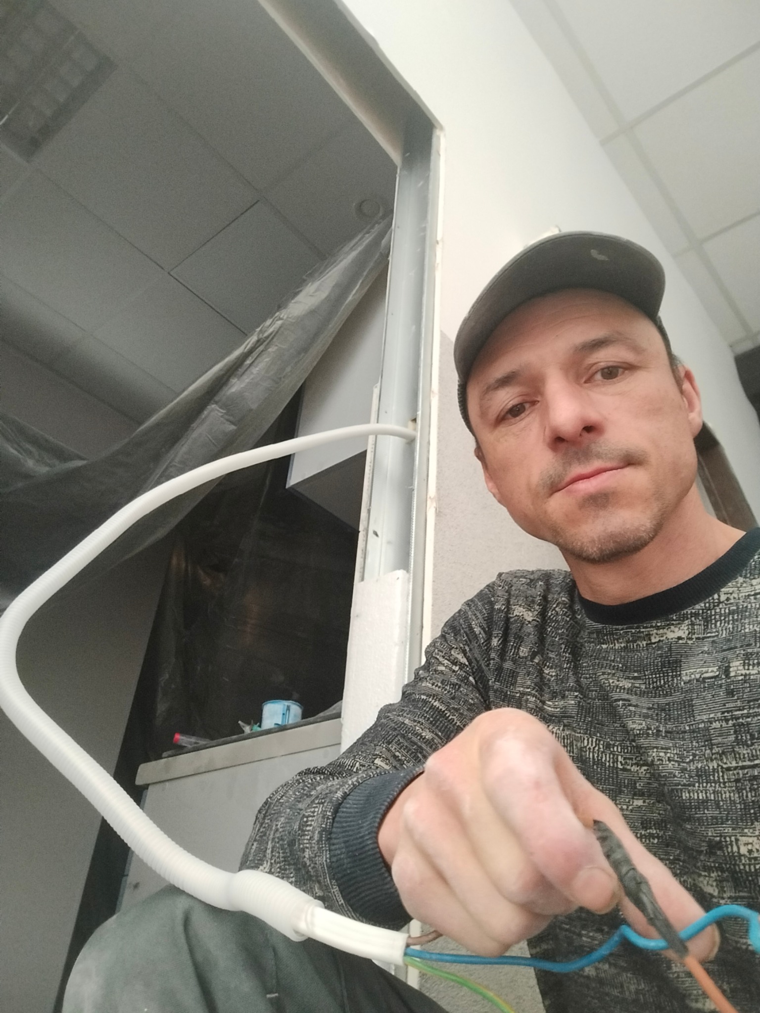 Elektryk w czapce, z bliska, trzyma przewody. W tle biała, pofałdowana rura instalacyjna i sufit podwieszany w trakcie montażu. Nietypowy kadr z góry.