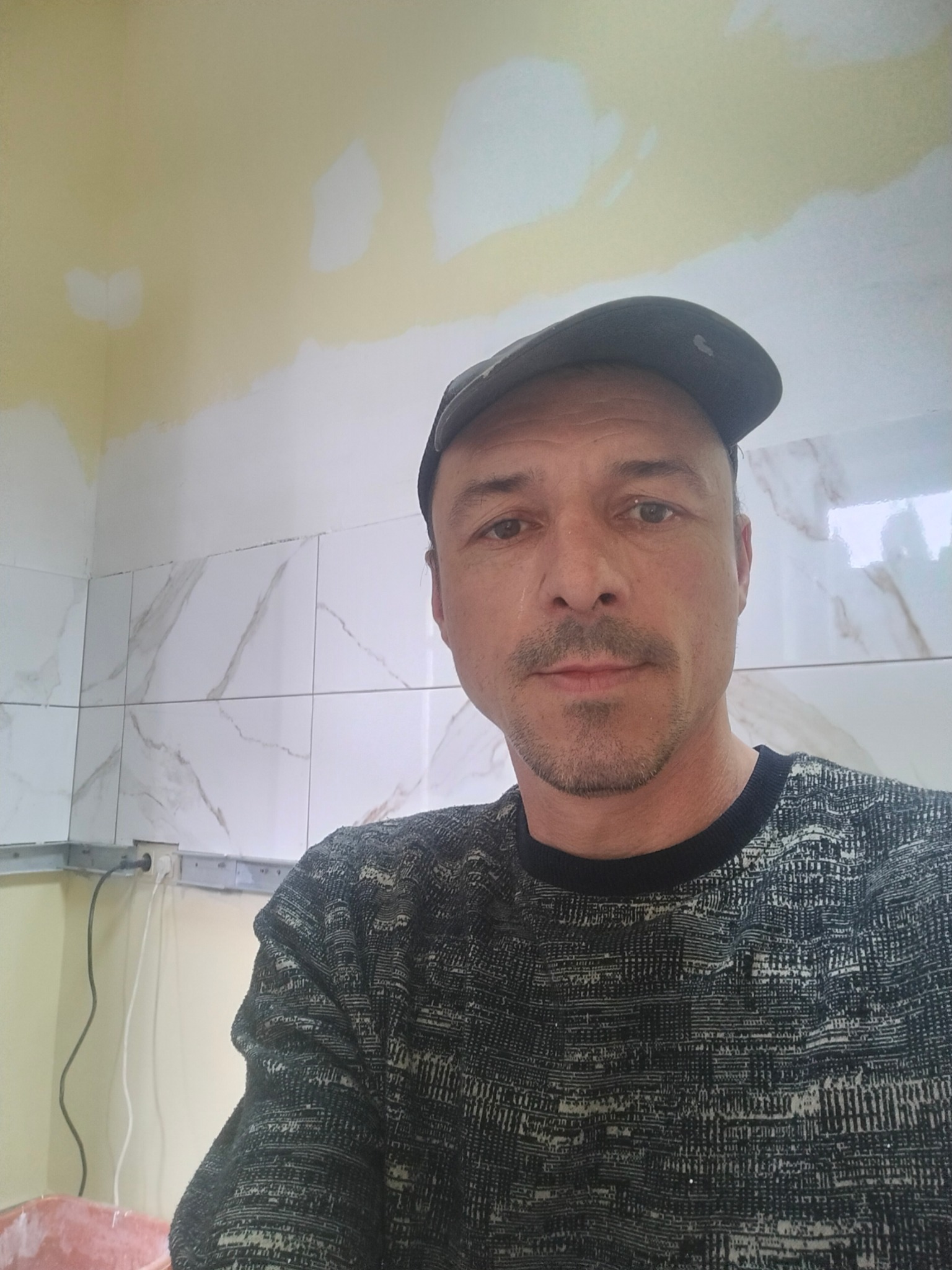 Mężczyzna w czapce na tle ściany z marmurowymi płytkami i żółtą, niedomalowaną górą w Trzebiatowie. Widoczne gniazdka i kable.