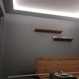 Coolwall Łukasz Rzepa - Szara sypialnia po remoncie: podświetlany sufit, drewniane półki i rama łóżka. Widoczny fragment otwartych drzwi. Świeżo pomalowane ściany.