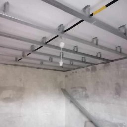 Coolwall Łukasz Rzepa - Metalowa konstrukcja sufitu podwieszanego z płyt karton-gips, widok z dołu na szkielet przed montażem płyt, surowe ściany w tle.