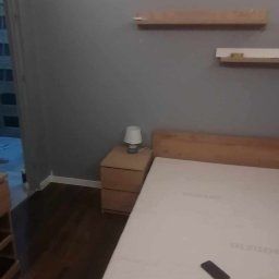 Coolwall Łukasz Rzepa - Sypialnia po remoncie: drewniane półki na szarej ścianie, podświetlany sufit, drewniane łóżko z białą pościelą i stolik nocny z lampką. Ciemna podłoga.