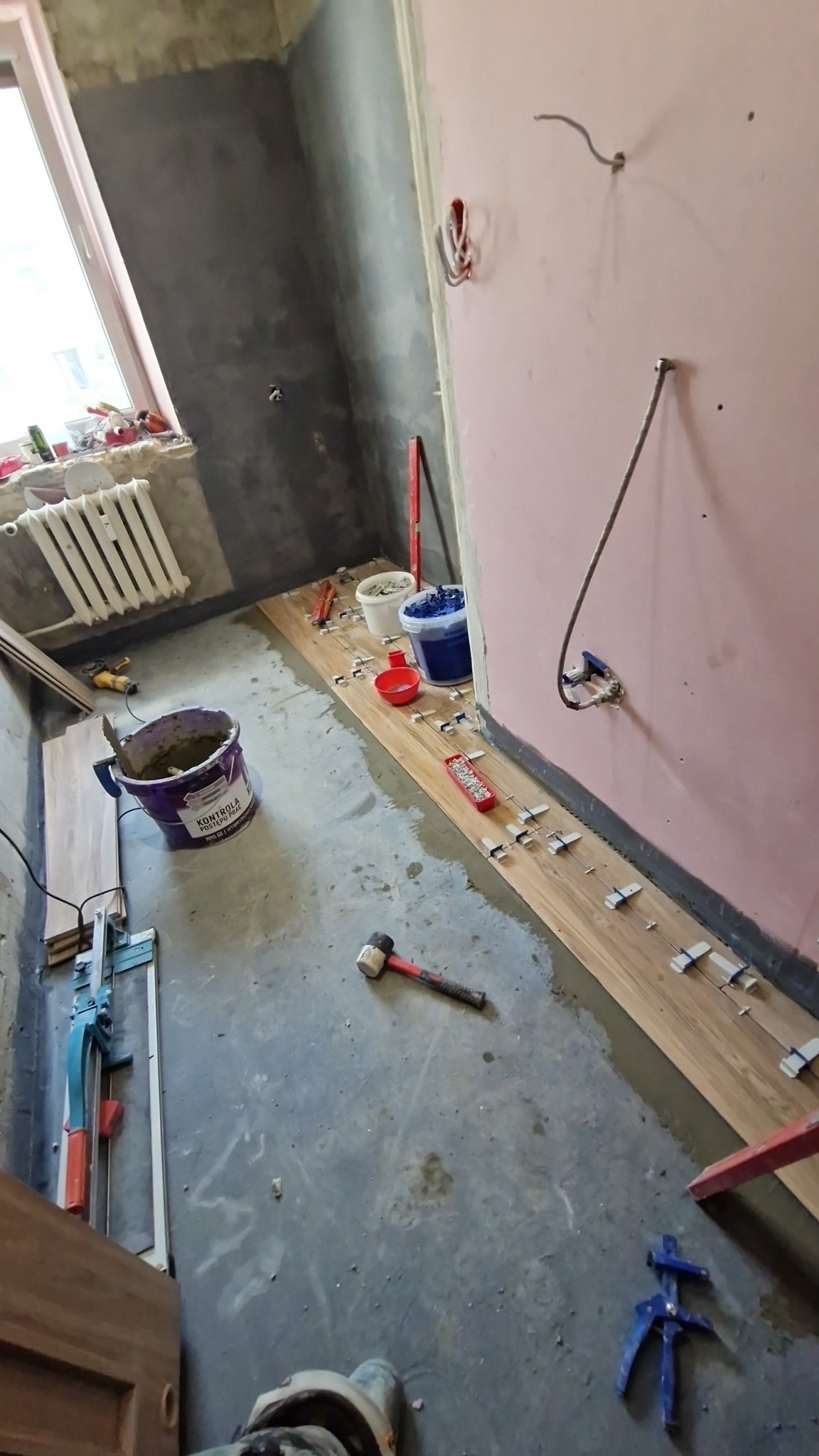 Układanie płytek imitujących drewno w trakcie remontu. Widoczne narzędzia glazurnicze, wiadro z zaprawą i fragmenty płytek. Ściany w surowym stanie, okno z grzejnikiem.