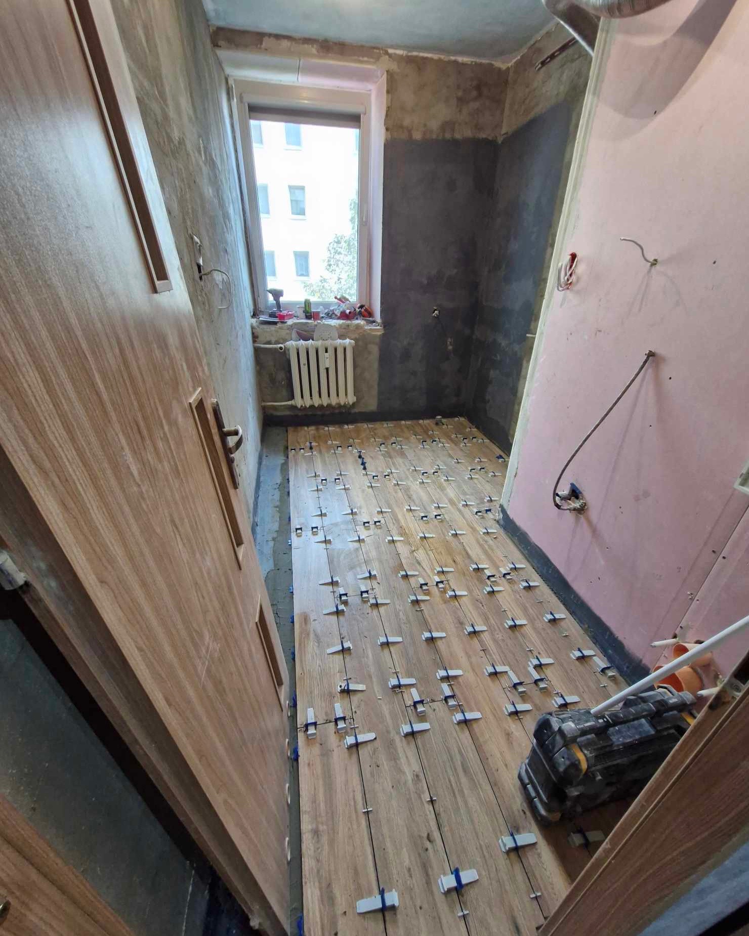 Układanie drewnopodobnych płytek podłogowych w trakcie remontu. Widoczne krzyżyki dystansowe, surowe ściany i okno z grzejnikiem.